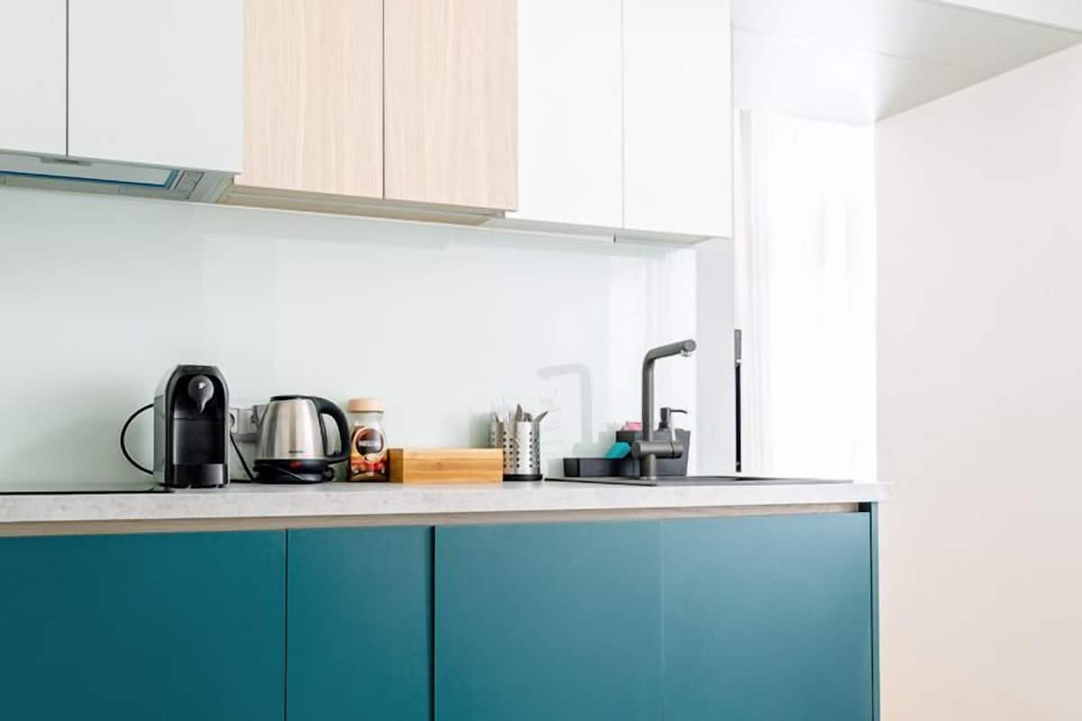 A modern kitchen with teal cabinetry, a wooden countertop, and a stainless steel kettle.