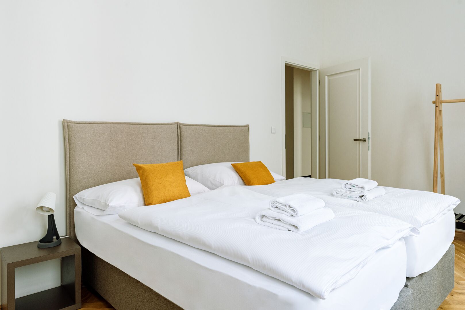 A double bed with white linens and two mustard pillows, a bedside table with a lamp, and a wooden floor.