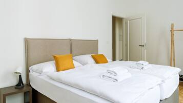 A double bed with white linens and two mustard pillows, a bedside table with a lamp, and a wooden floor.