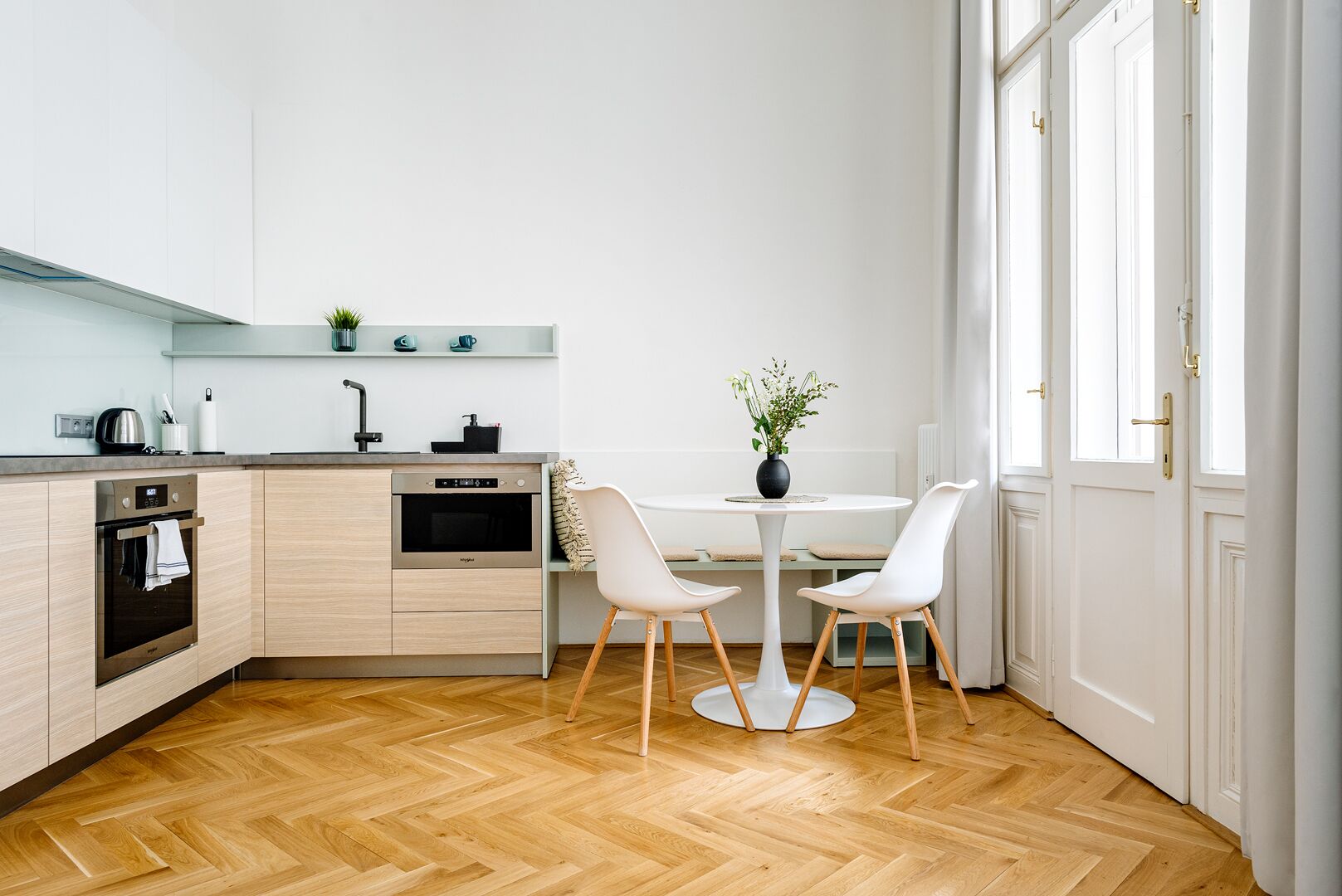 A modern kitchen with a dining area, wooden herringbone flooring, and a large window.