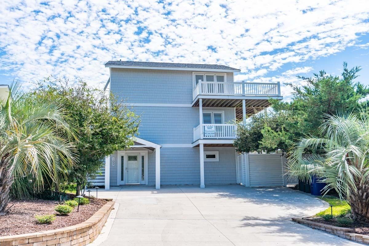 A two-story residential building with a balcony on the upper floor, surrounded by landscaped areas and palm trees.