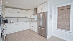 A modern kitchen with white cabinetry, a stainless steel refrigerator, and a window with a bamboo blind. - Emerald Isle Lookout, Oceanview House in Emerald Isle, NC! (Emerald Isle)