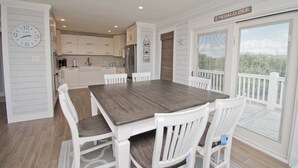 A dining area with a wooden table and white chairs, a kitchen in the background, and a sign reading "Emerald Isle". - Emerald Isle Lookout, Oceanview House in Emerald Isle, NC! (Emerald Isle)