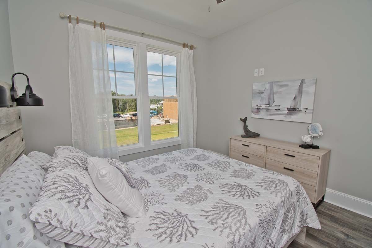 Un dormitorio con cama, ventana con cortinas, mesita de noche, lámpara y un cuadro en la pared.