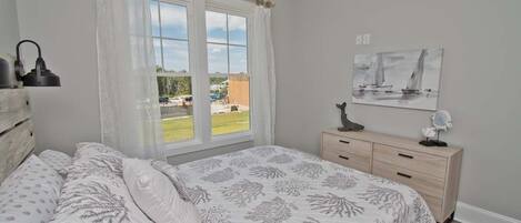 Un dormitorio con cama, ventana con cortinas, mesita de noche, lámpara y un cuadro en la pared.
