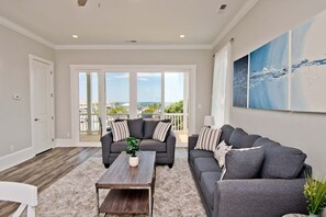 A spacious living room with a gray sofa, a wooden coffee table, and a large window offering a view of the ocean. - Village West #16, Oceanview Townhouse in Emerald Isle, NC! (Emerald Isle)