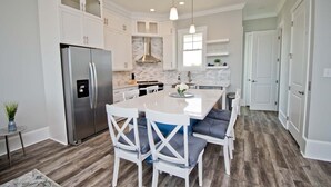 A modern kitchen with white cabinetry, a stainless steel refrigerator, a central island with seating, and a herringbone-patterned backsplash. - Village West #16, Oceanview Townhouse in Emerald Isle, NC! (Emerald Isle)