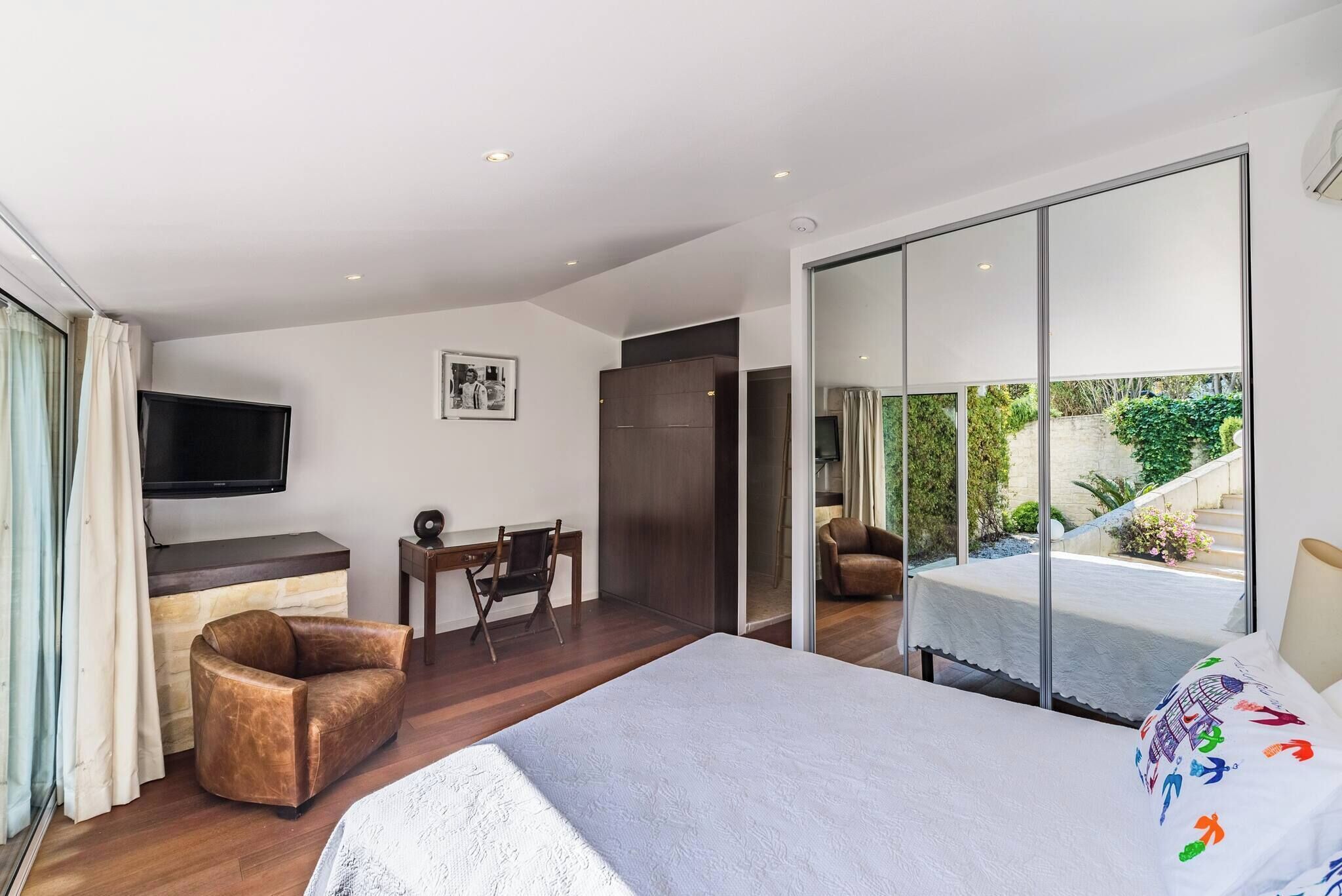 Camera d'albergo moderna con un letto, una scrivania, una sedia, una televisione e una porta scorrevole in vetro che dà su un balcone con vista sul verde.