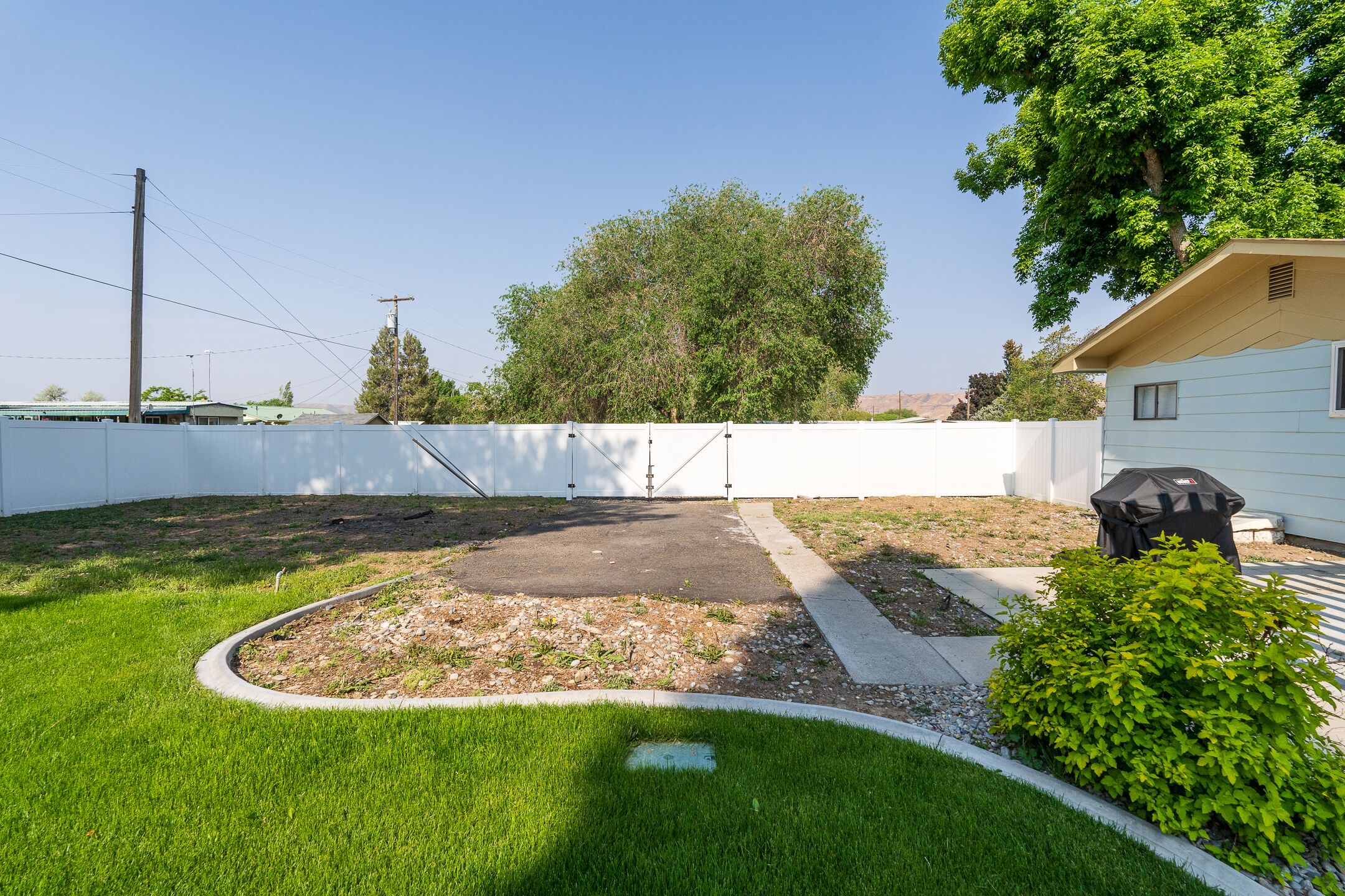 Un jardin à l’arrière d’une maison, avec une zone dallée, une clôture blanche et un petit arbre.