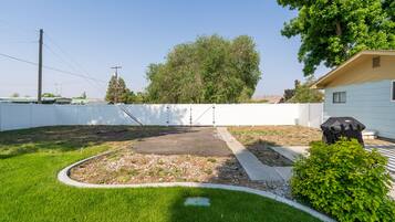Un jardin à l’arrière d’une maison, avec une zone dallée, une clôture blanche et un petit arbre.