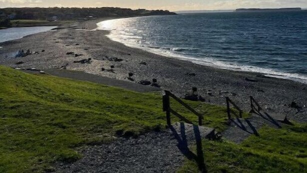 Paesaggio costiero con cielo sereno, spiaggia illuminata dal sole e area erbosa con recinzione in legno.