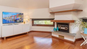 A modern living room with a wooden floor, a flat-screen TV mounted on a white cabinet, a fireplace with a stone mantle, and a window with a view of greenery.