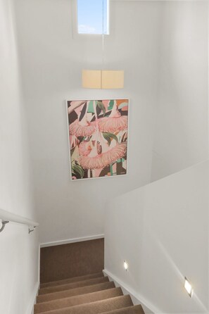 Una escalera con un cuadro de flores rosadas colgado en la pared, una lámpara de techo y una ventana.