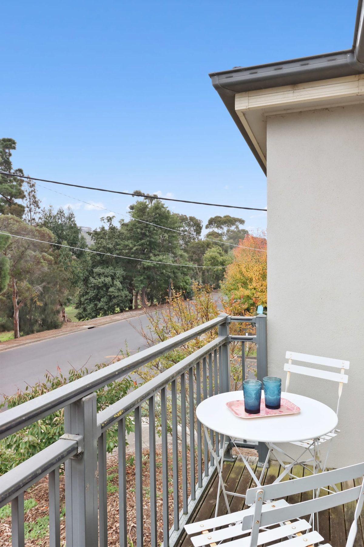 Ein Balkon mit einem weißen Tisch und Stühlen, der auf eine Straße und Bäume blickt.