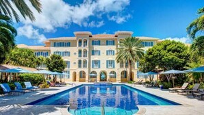 A large pool with lounge chairs and umbrellas, surrounded by a building with multiple balconies and a clear blue sky. - Villa Renaissance 2 Story, 4 Bedrooms, 4 1/2 Baths, Ocean Front Master Bedroom (Grace Bay)