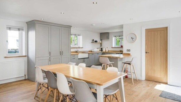 A modern kitchen with white cabinets, a wooden dining table, and a clock on the wall. - Modern Home in Bude, Private Parking & Garden (Bude)