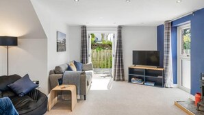 A living room with a sofa, a coffee table, a TV on a stand, and a view through a door to an outdoor area. - Modern Home in Bude, Private Parking & Garden (Bude)