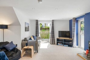 A living room with a sofa, a coffee table, a TV on a stand, and a view through a door to an outdoor area. - Modern Home in Bude, Private Parking & Garden (Bude)