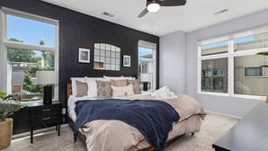 Quarto com cama, mesas de cabeceira, ventilador de teto e grandes janelas.