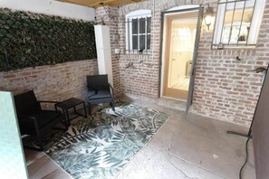 A room with a brick wall, a window with bars, a door, a black chair, a small table, and a patterned rug.