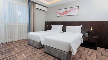 Een hotelkamer met twee bedden, een nachtkastje met een telefoon, een schilderij aan de muur en een raam met dunne gordijnen.