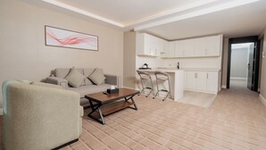 Una sala de estar moderna con un sofá, una mesa de centro y un sillón estampado. Se observa una zona de cocina con armarios blancos y un taburete de bar.