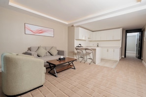 Una sala de estar moderna con un sofá, una mesa de centro y un sillón estampado. Se observa una zona de cocina con armarios blancos y un taburete de bar.
