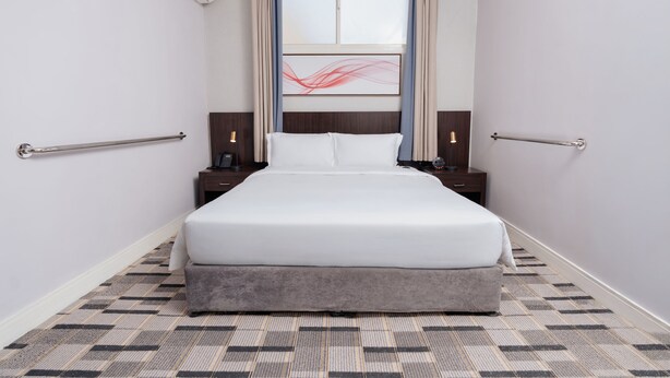 Una habitación de hotel con cama, mesitas de noche, teléfono, lámpara y ventana con cortinas.