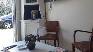 A room with a television mounted on the wall, a cabinet with glass doors, a table with a vase of flowers, and a chair. - Maison 70m2 (L'Épine)