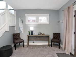 Um quarto com uma parede cinzenta, um banco de madeira, duas cadeiras castanhas, uma mesinha com objetos decorativos, uma janela e uma porta com cortinas.