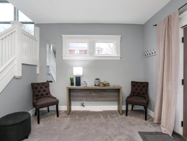 Una habitación con pared gris, un banco de madera, dos sillas marrones, una mesa pequeña con adornos, una ventana y una puerta con cortinas.