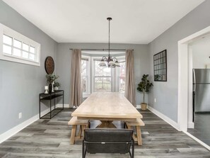 Un comedor con una gran mesa de madera, bancos, una lámpara colgante y vistas amplias a través de ventanas grandes.