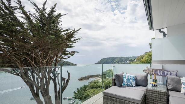 A balcony with wicker furniture, a glass railing, and a view of the sea and hills. - Blackstone -  stylish apartment with incredible views looking out over the Salcombe estuary (Salcombe)