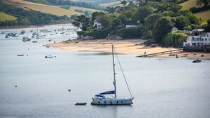 A coastal scene with a sailboat anchored near a sandy beach, surrounded by hills and residential buildings. - Blackstone -  stylish apartment with incredible views looking out over the Salcombe estuary (Salcombe)