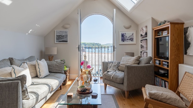 Un salon chaleureux avec une table basse en verre, deux canapés, une bibliothèque et une vue sur la mer par une porte ouverte.