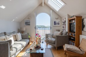 Ett mysigt vardagsrum med ett soffbord i glas, två soffor, en bokhylla och utsikt över havet genom en öppen dörr.