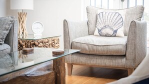 Un salon moderne avec un fauteuil gris agrémenté d’un coussin en forme de coquillage, une table basse en verre, une table d’appoint en bois et une lampe.