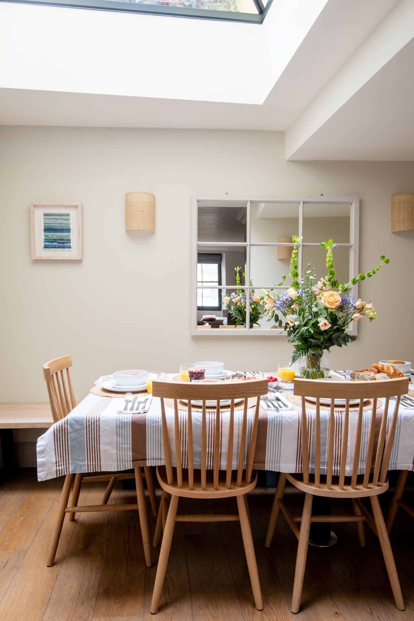 A dining area with a table set for four, wooden chairs, a vase of flowers, and a mirror on the wall.