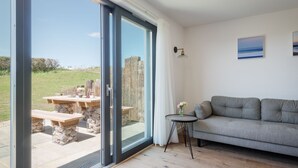 Una sala de estar moderna con un sofá gris, una mesita redonda con un jarrón de flores, y un amplio ventanal corredero que da a un área exterior con una mesa y sillas de madera.