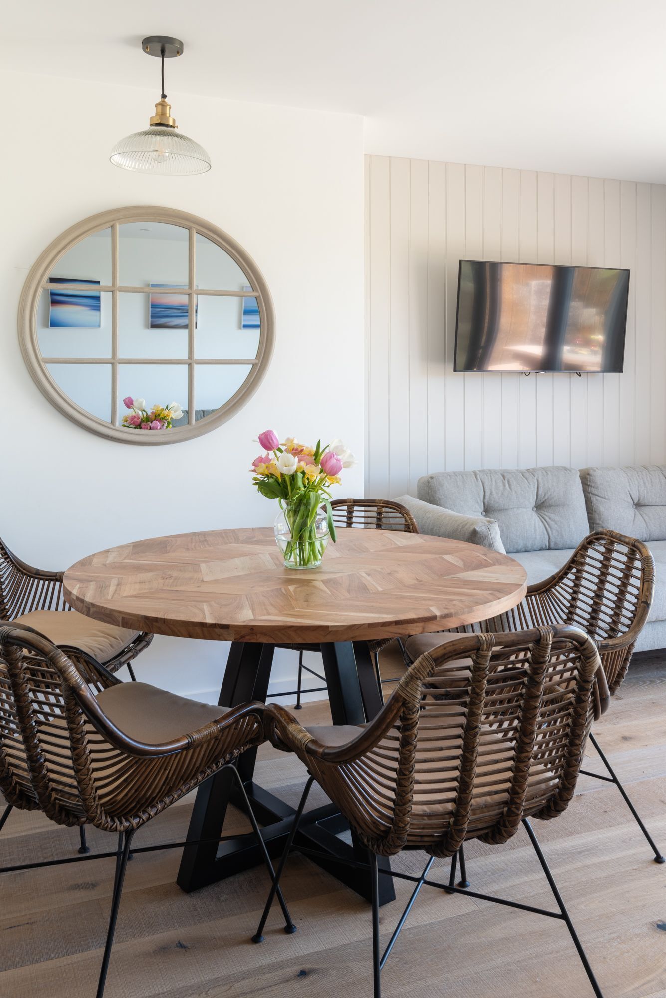 Une table de salle à manger ronde en bois, entourée de six chaises en osier, un vase de fleurs, un miroir rond et un téléviseur fixé au mur.