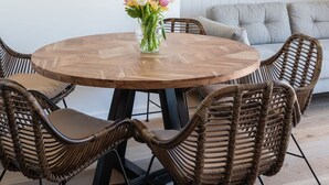 Una mesa de comedor redonda de madera con seis sillas de mimbre, un jarrón de flores, un espejo redondo y un televisor montado en la pared.