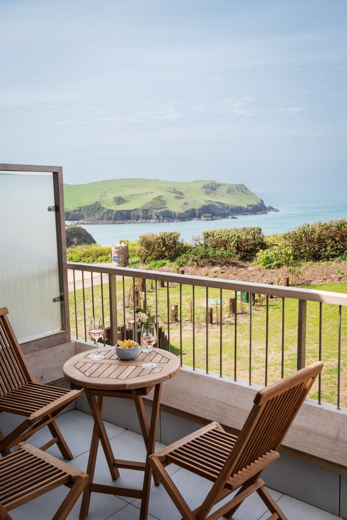 A balcony with a wooden table and chairs, overlooking a coastal landscape with hills and the sea.