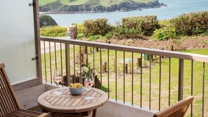 A balcony with a wooden table and chairs, overlooking a coastal landscape with hills and the sea. - Searush Cove - a beautiful contemporary beachfront apartment (Hope Cove)