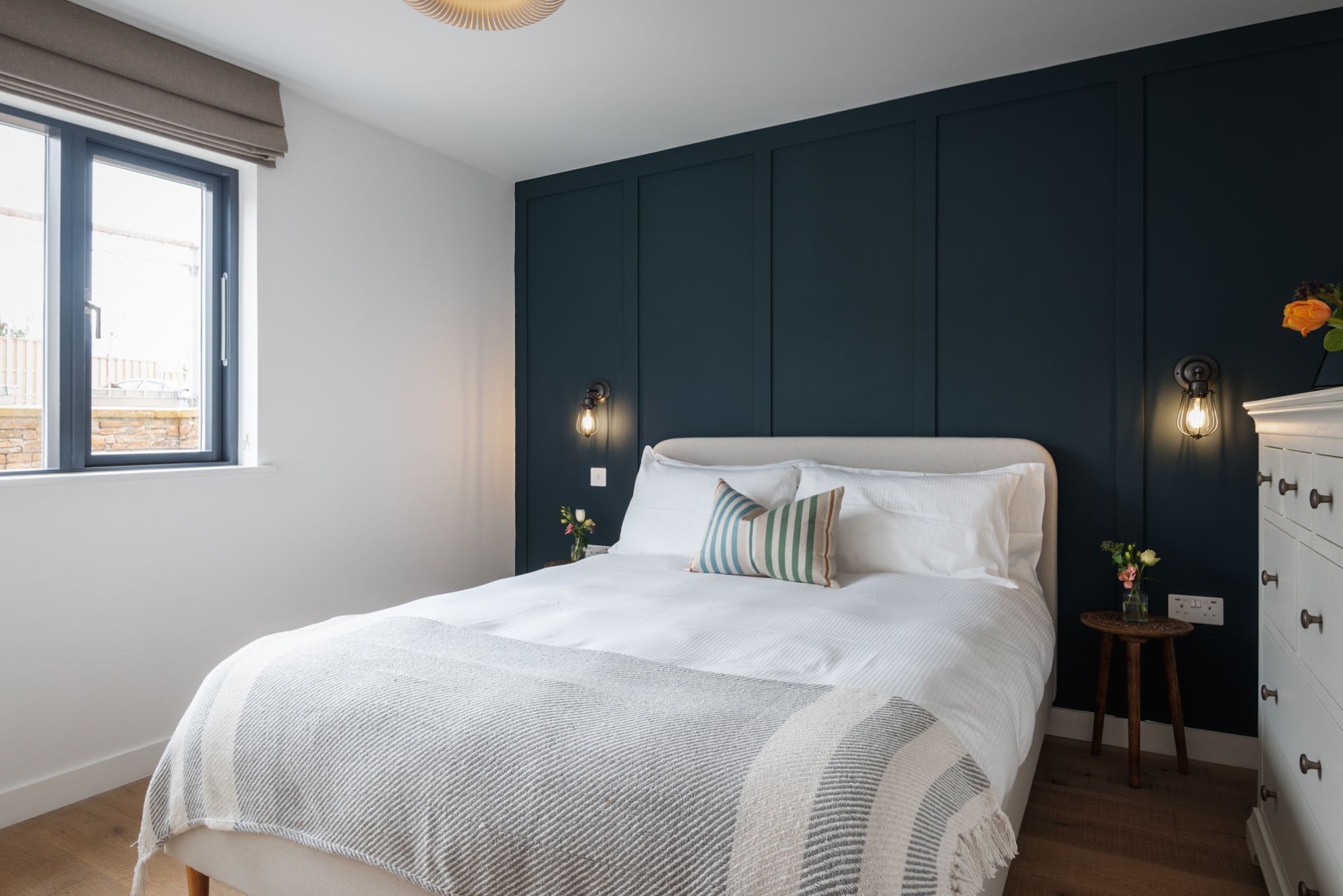 A bedroom with a large bed, two wall sconces, a dresser, and a window with a view.