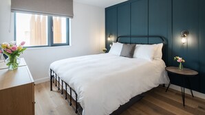 A bedroom with a bed, a bedside table, a nightstand, a window with curtains, and a wall-mounted light fixture. - Willow Cove -  a luxury self-catering apartment footsteps from the beach (Hope Cove)