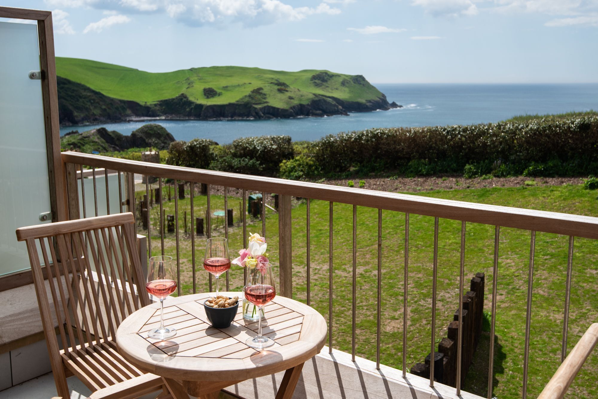 Ein Balkon mit Tisch für zwei Personen, Blick auf eine Küstenlandschaft mit grünen Hügeln und dem Meer.