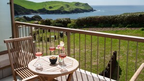 A balcony with a table set for two, overlooking a coastal landscape with green hills and the sea. - Willow Cove -  a luxury self-catering apartment footsteps from the beach (Hope Cove)