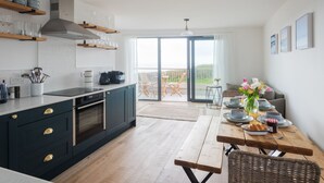 A modern kitchen with dark cabinetry, a built-in oven, and a coffee maker. There is a dining area with a wooden table, chairs, and a view of the outdoors through large sliding doors. - Willow Cove -  a luxury self-catering apartment footsteps from the beach (Hope Cove)