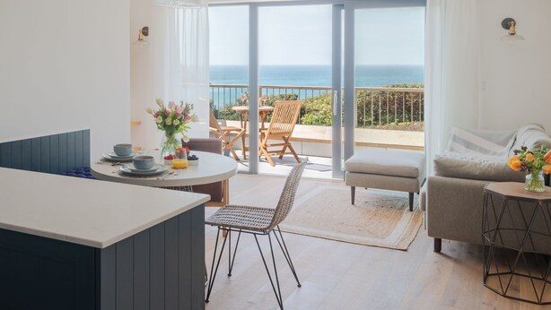 A modern living room with a view of the sea, a dining area with a table set for two, and a cozy seating area with a sofa and armchair.