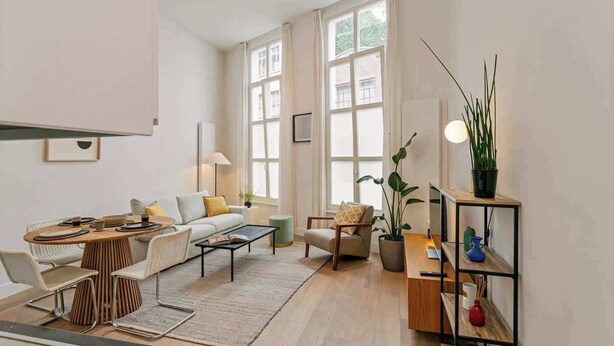 Ein modernes Wohnzimmer mit Sofa, Couchtisch und Essbereich. Große Fenster, eine Pflanze und ein Bücherregal sind ebenfalls vorhanden.